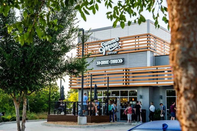 Modern coffee shop with horizontal wood and metal slatted facade, customers gathered outside entrance, surrounded by trees