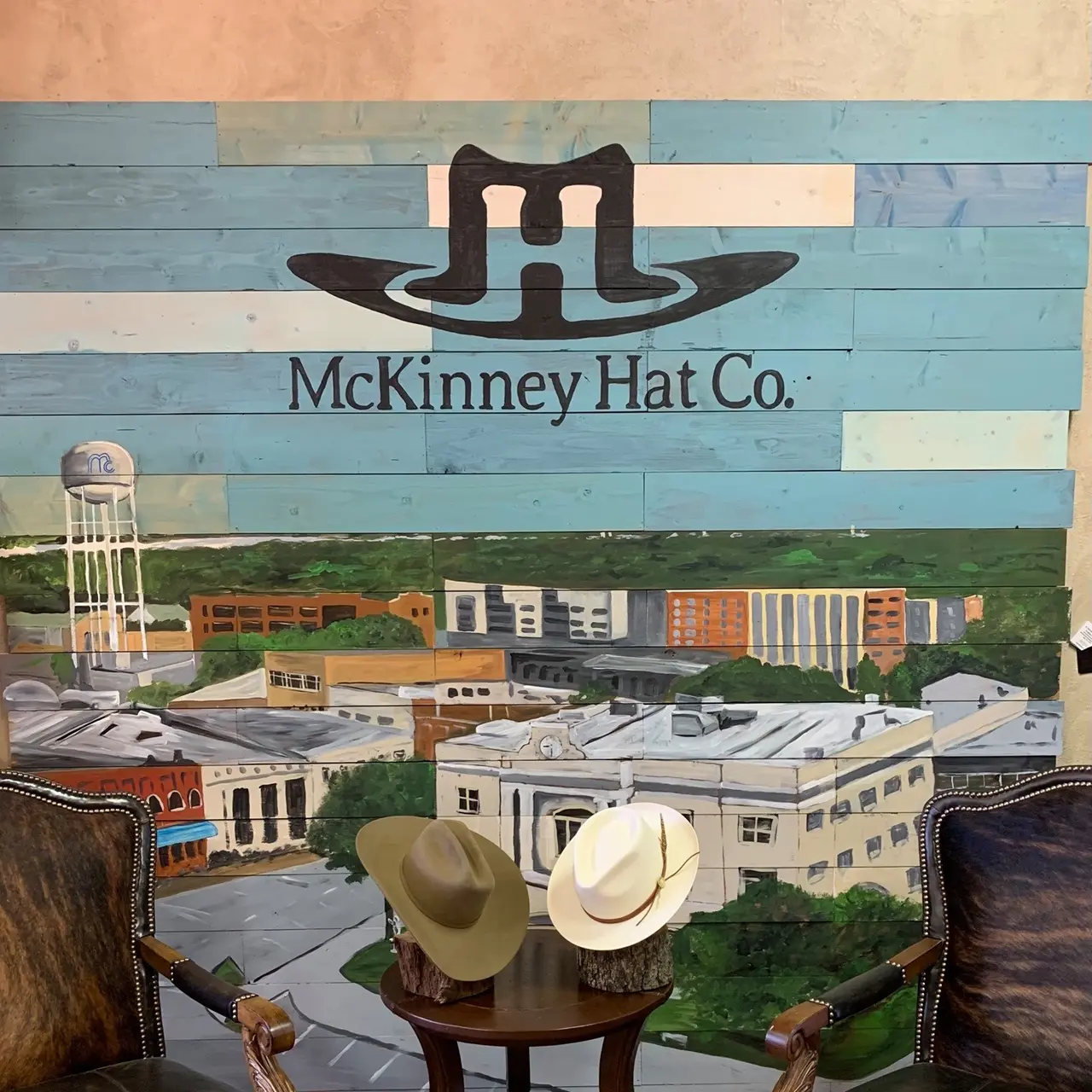 Mural with stylized hat logo and downtown skyline featuring two cowboy hats displayed on wooden stand in foreground