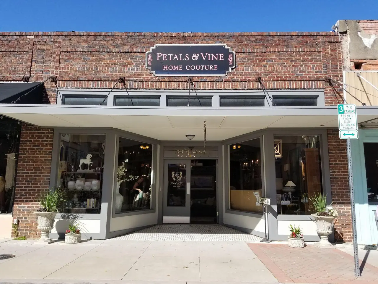 Brick storefront with large windows displaying home décor items, potted plants flanking the entrance doors