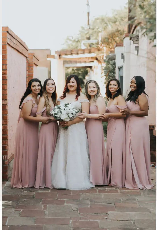 Bride in white gown with five bridesmaids in matching pink dresses holding white flowers in brick courtyard