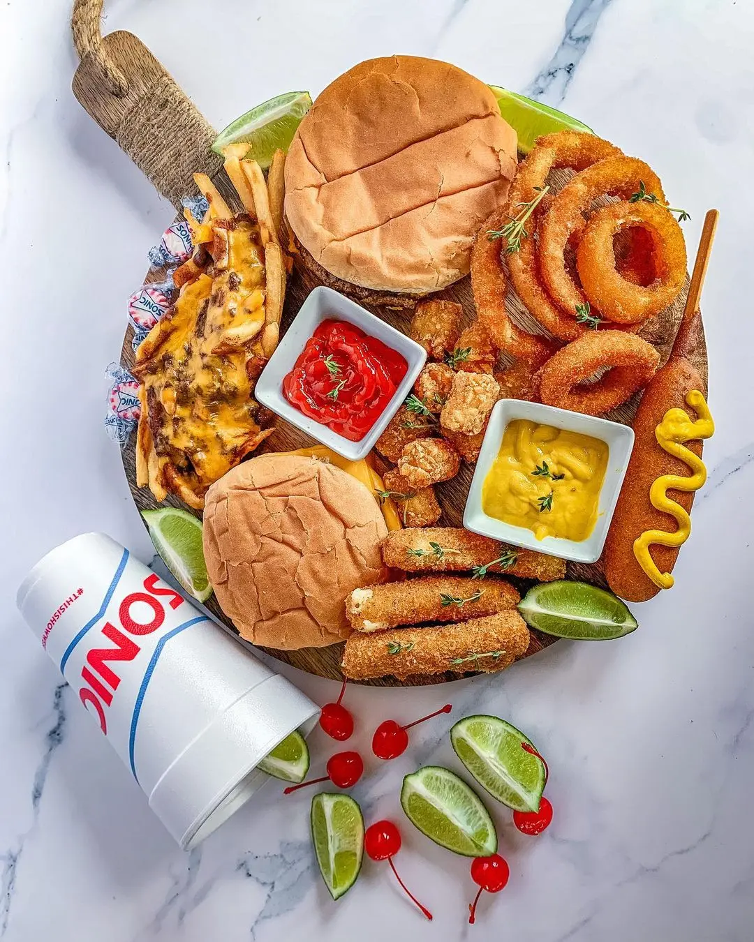 Wooden serving board with two burgers, onion rings, fried chicken tenders, corn dog, and small bowls of ketchup and mustard, alongside a drink cup and lime