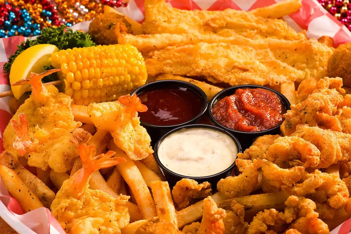 Fried seafood platter with corn, shrimp, fish, and onion rings arranged around three sauce bowls on checkered paper