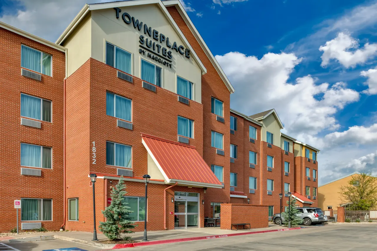 Brick hotel building with pitched roof, ground-level entrance with red awning, multiple windows, and parking area under blue sky