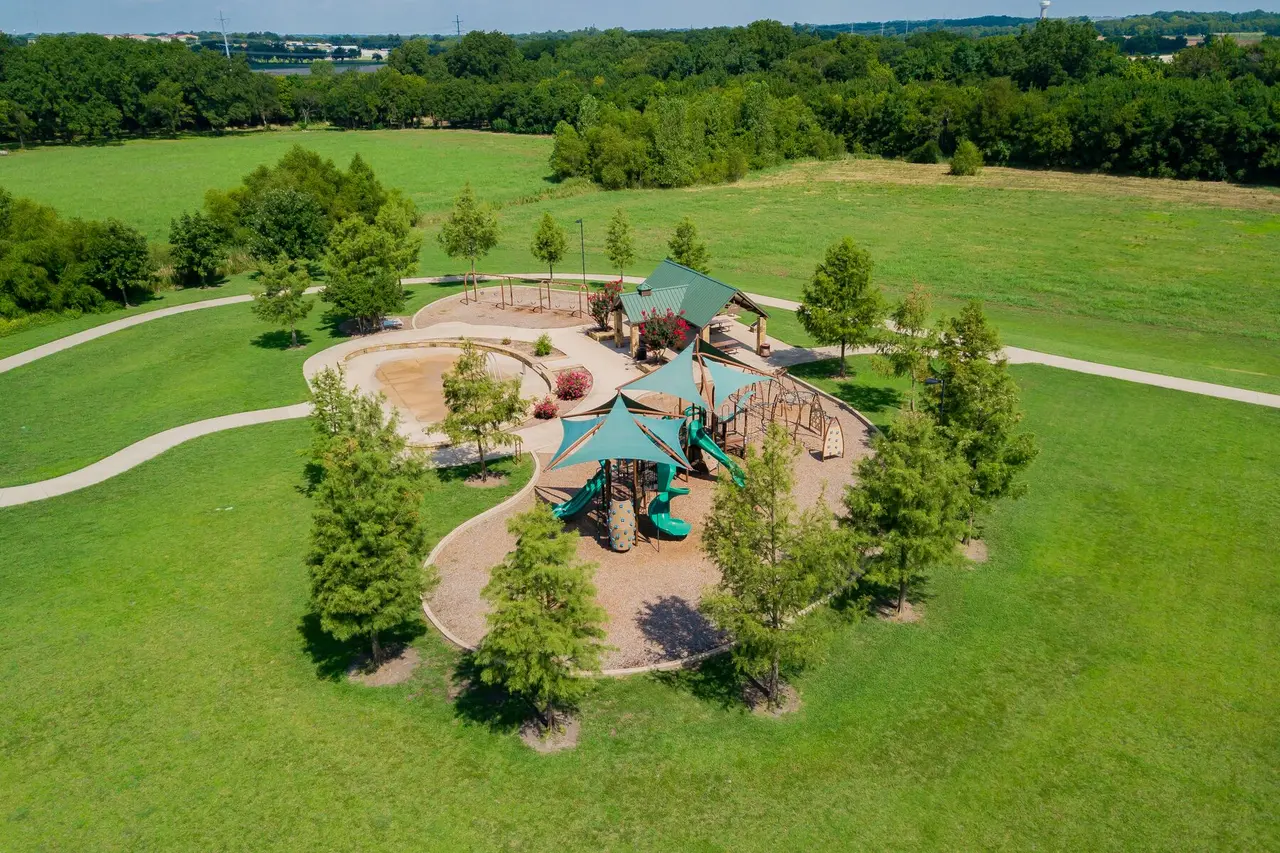 Aerial view of a neighborhood park with playground structures, splash pad, and surrounding trees on green lawn