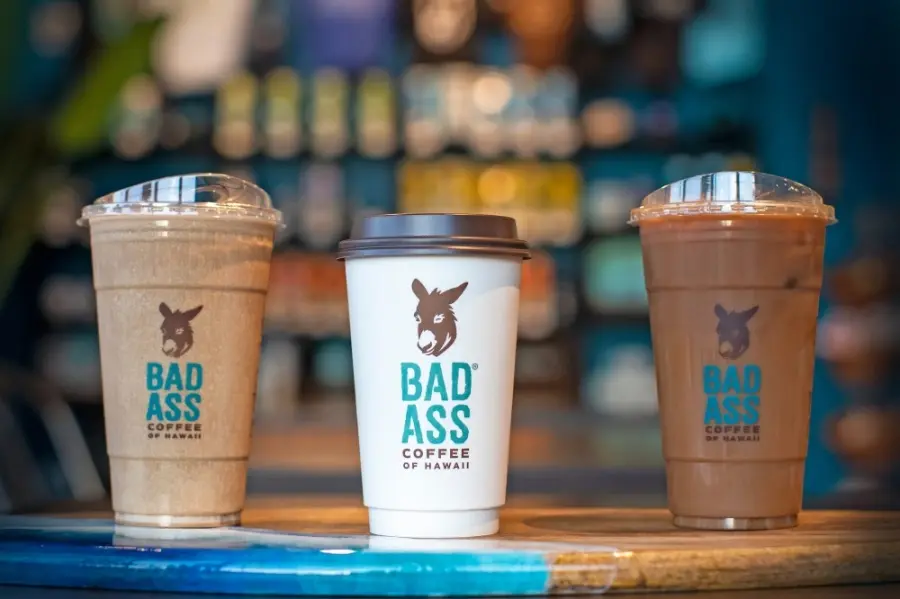 Three coffee drinks with Bad Ass Coffee branding on a blue counter, with a blurred cafe interior in background