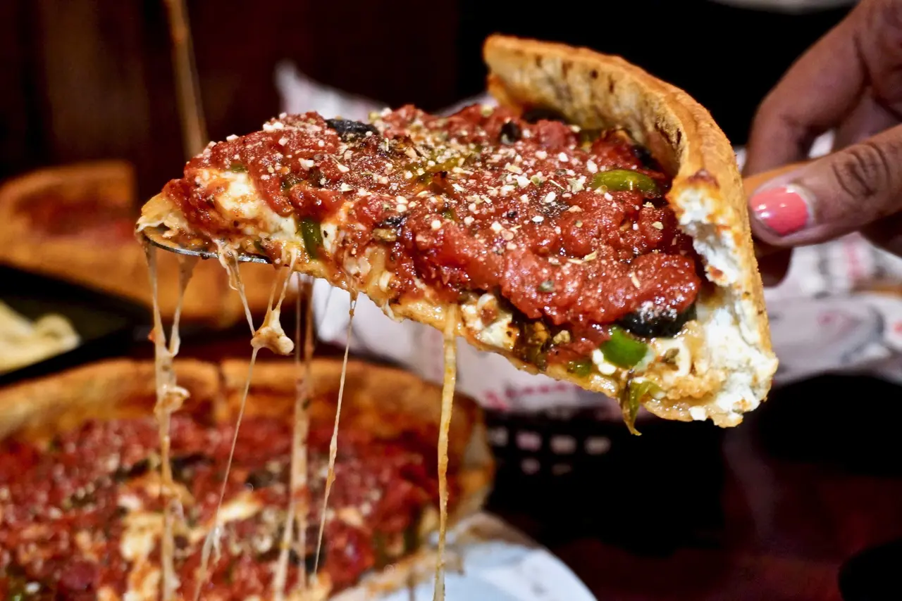 Hand holding a slice of deep-dish pizza with melted cheese stretching, topped with tomato sauce and herbs