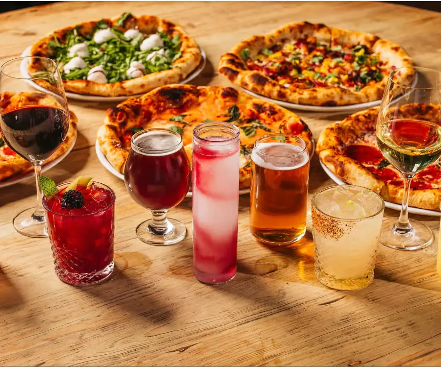 Pizzas and cocktails arranged on wooden table, including red and pink beverages in glasses alongside wine and beer