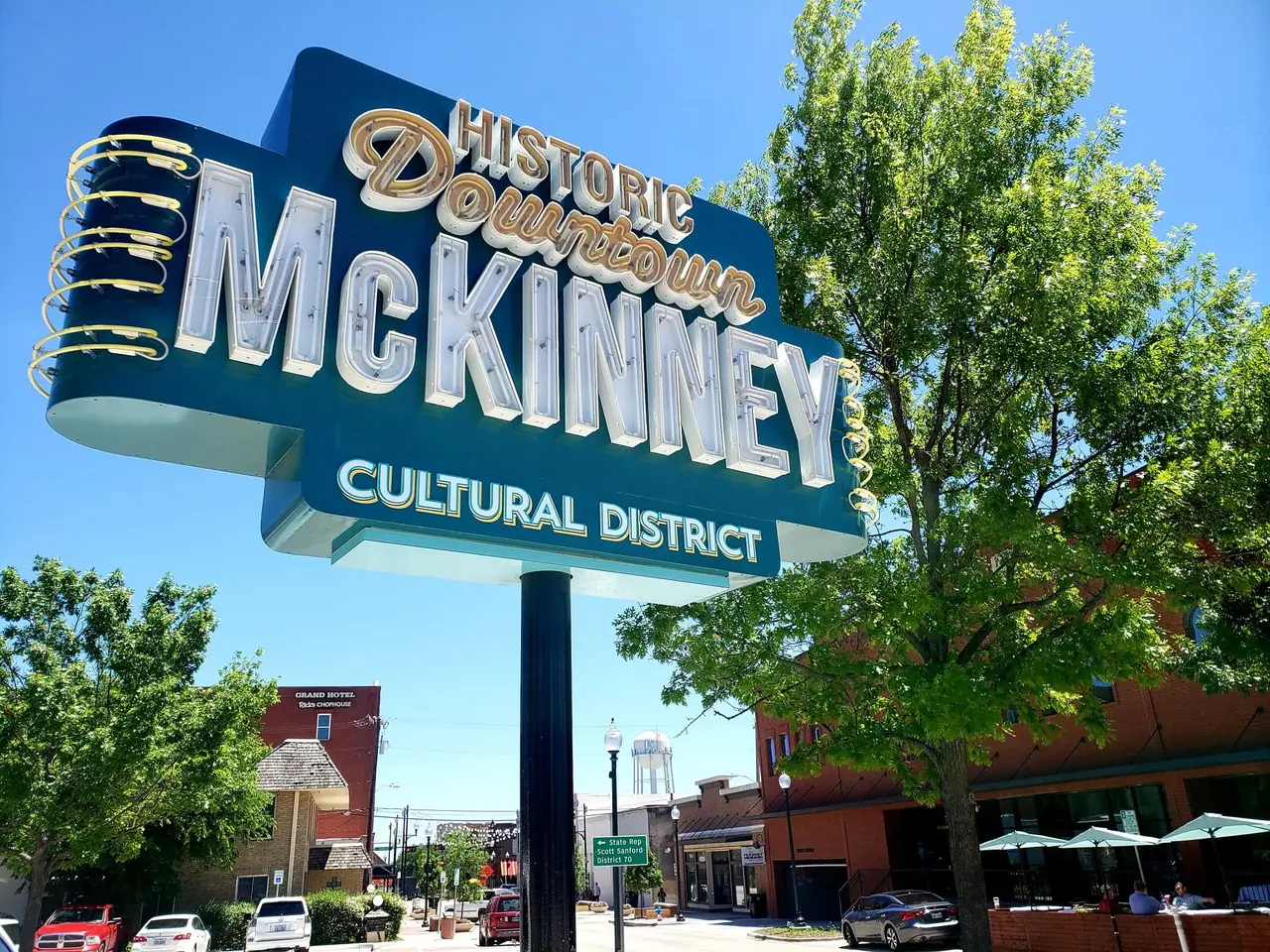 Large vintage-style sign reading "Historic Downtown McKinney Cultural District" with tree-lined street and brick buildings below