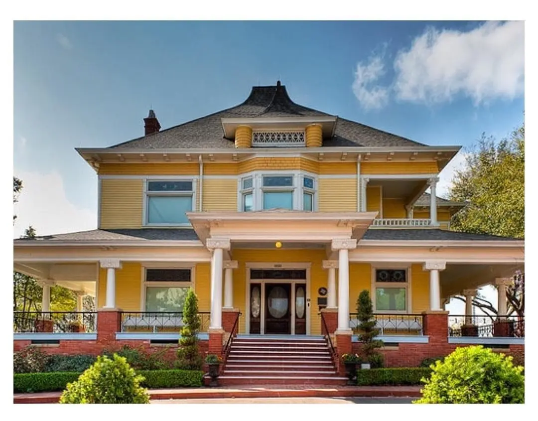 Victorian-era mansion with yellow exterior, red brick base, wrap-around porch, and mansard roof under blue sky