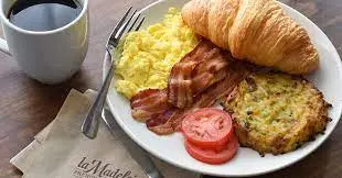 Breakfast plate with scrambled eggs, bacon, croissant, hash brown, tomato slices, and cup of coffee