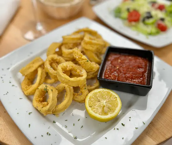 Fried calamari rings on white plate with lemon slice and red dipping sauce, salad visible in background