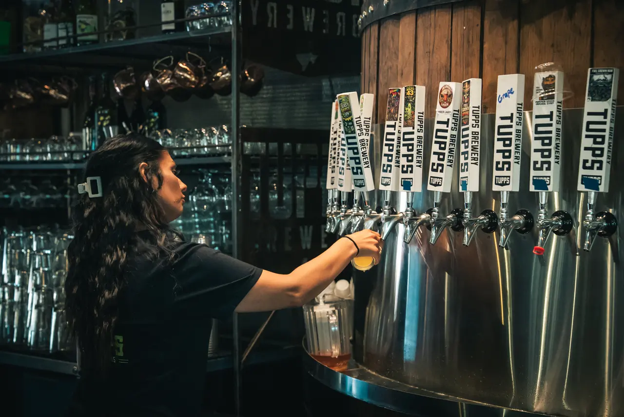 Person pouring beer from a tap at a brewery bar, multiple labeled taps visible above
