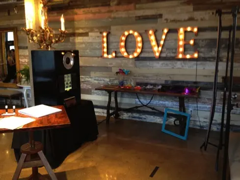 Event venue interior with illuminated LOVE sign, photo booth setup, and decorated table against wooden wall