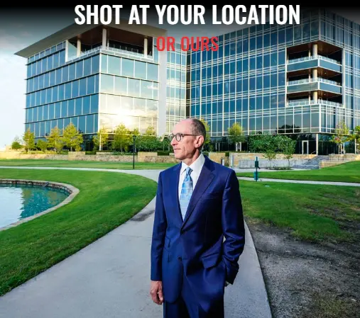 Man in dark suit and glasses standing on a paved path in front of a modern glass office building with manicured lawn and water feature