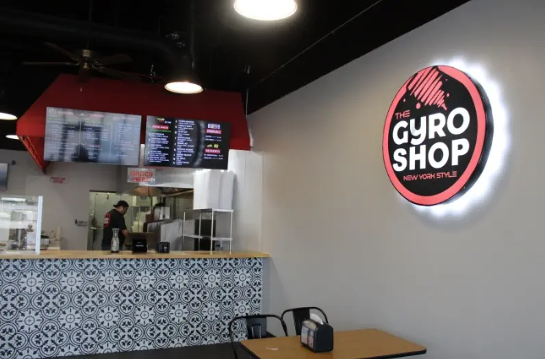 Interior counter with neon Gyro Shop sign, menu board above service area, and wooden table with chairs in foreground