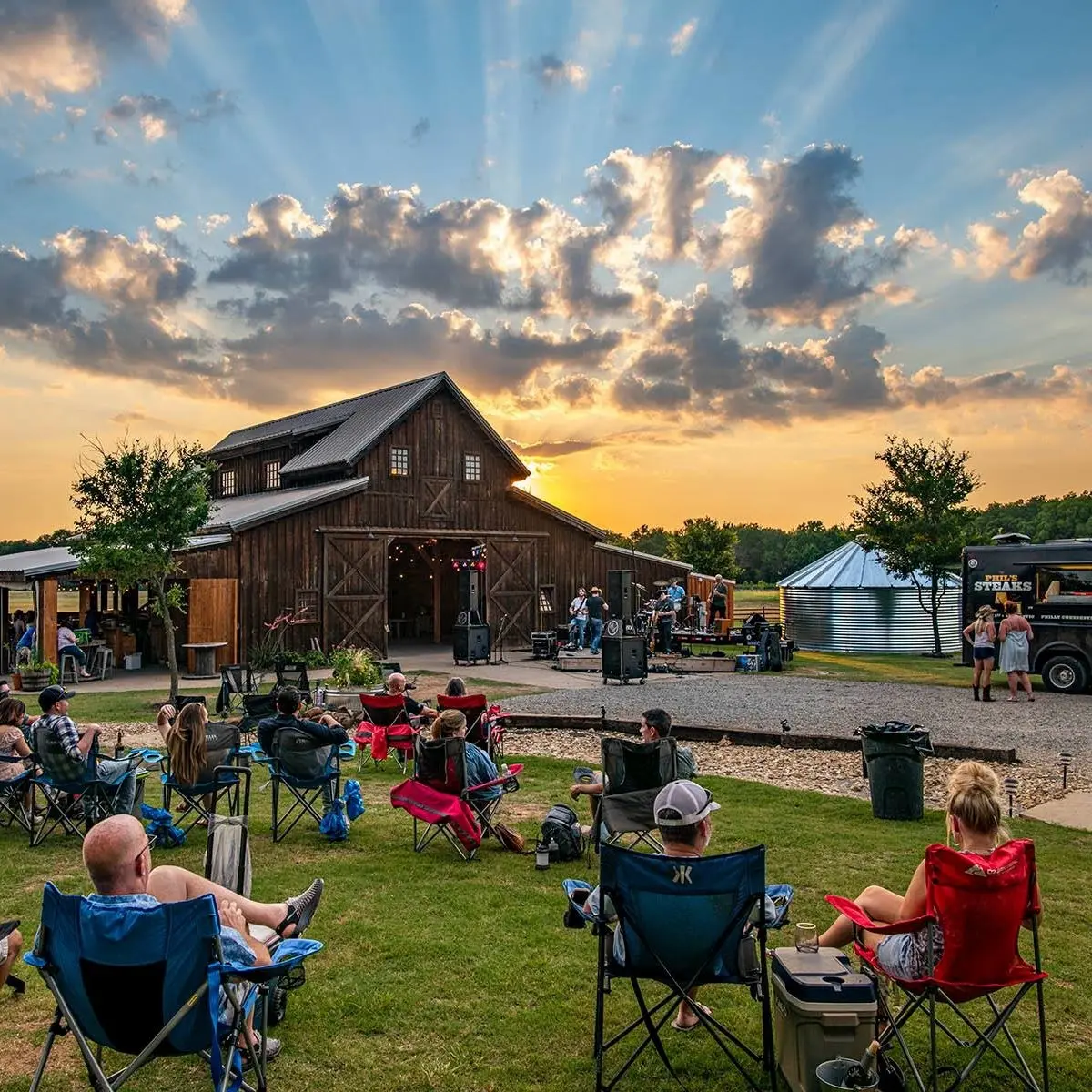 Visitors seated on lawn chairs watching sunset over a wooden barn with people gathered outside, trees and farm structures visible