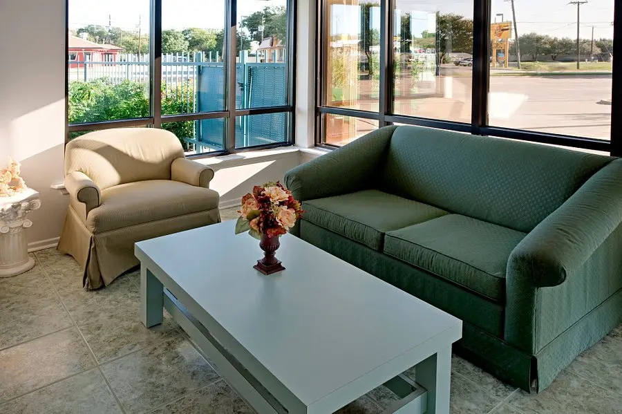 Bright lobby lounge with beige armchair, green sofa, white coffee table, and flower arrangement, viewed through large windows to parking area