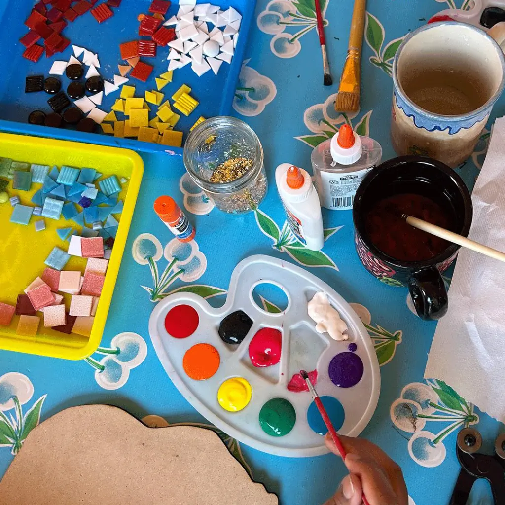 Art supplies and materials arranged on a blue table including a paint palette, colored paper squares, brushes, paint containers, and decorative items