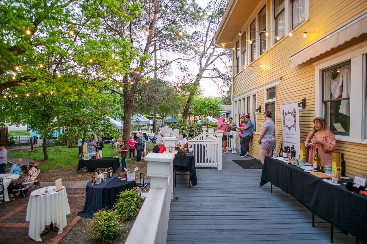 Outdoor event on deck and grounds of historic home with attendees mingling, tables with food and drinks, surrounded by mature trees