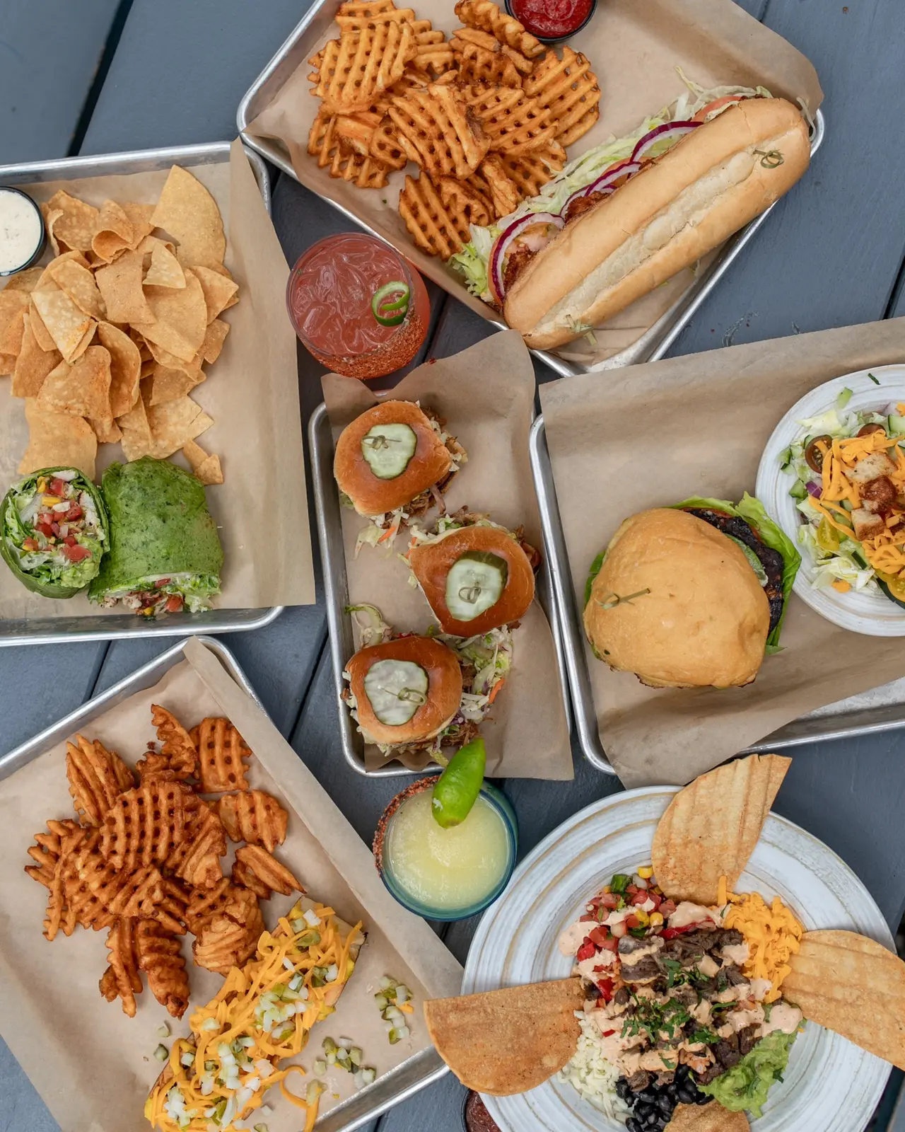 Overhead view of food in paper containers and plates: waffle fries, hot dog with toppings, chips, sandwiches, burgers, and salads on a gray surface