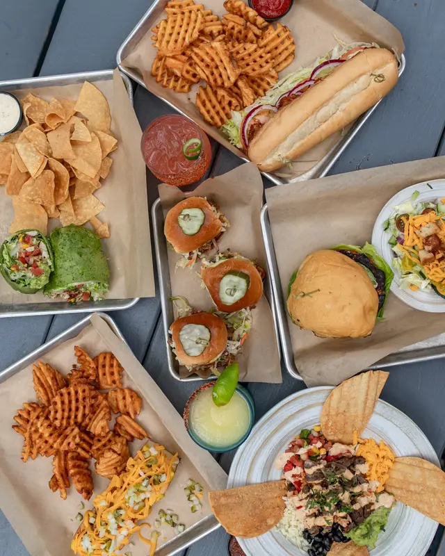 Overhead view of food in paper containers and plates: waffle fries, hot dog with toppings, chips, sandwiches, burgers, and salads on a gray surface