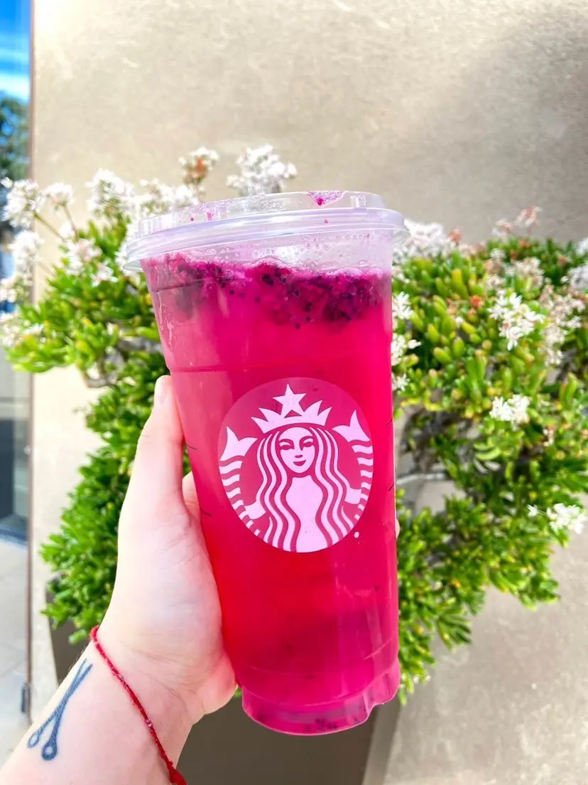 Hand holding a bright pink beverage in a clear cup with white logo, small white flowers visible in background