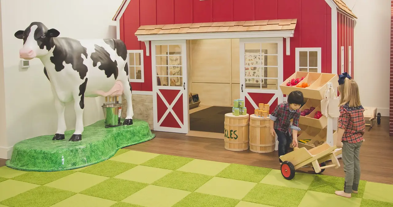Children playing around a toy farm barn with a large cow figure and farm equipment props