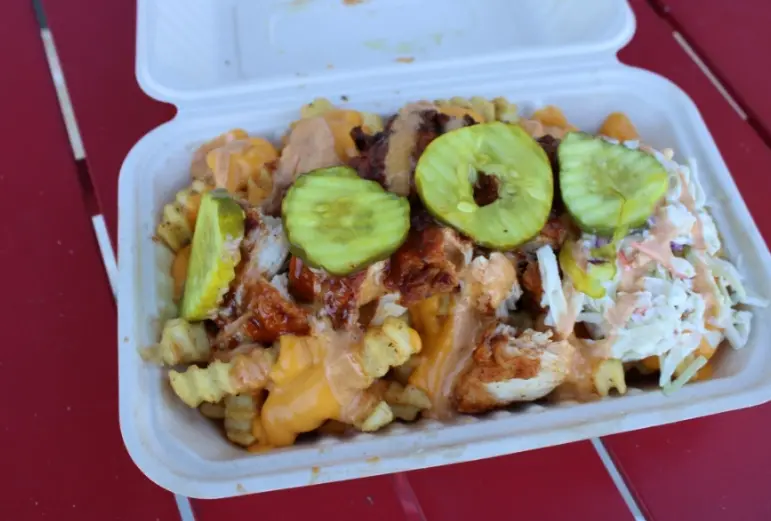 Loaded fries topped with shredded chicken, melted cheese, crispy bacon, and fresh cucumber slices in a white takeout container
