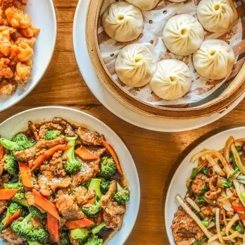 Bamboo steamer of dumplings, plate of stir-fried meat with broccoli and carrots, plate of crispy noodles with meat and vegetables, and plate of fried popcorn
