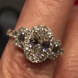Diamond engagement ring with oval-cut center stone and surrounding smaller diamonds on white metal band, worn on finger