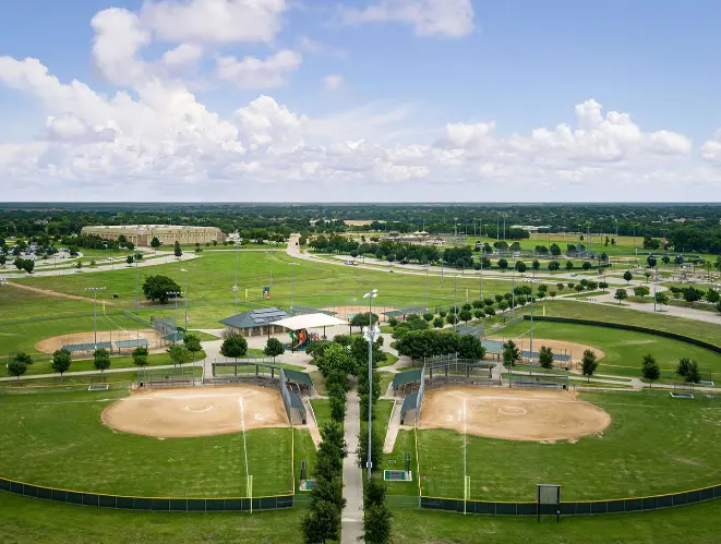 Aerial view of two lighted softball fields with dugouts surrounded by green grass and trees in a sports complex