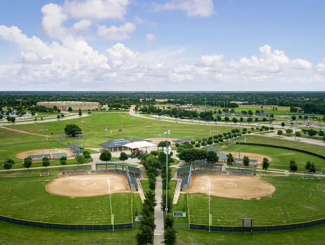Aerial view of two lighted softball fields with dugouts surrounded by green grass and trees in a sports complex