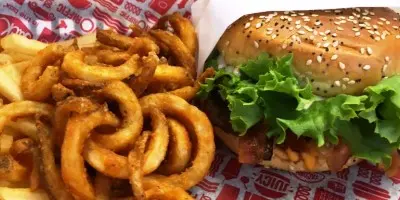 Burger with lettuce and sesame seed bun beside a heap of curly fried onion rings on checkered paper