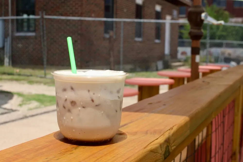 Iced coffee drink with green straw on wooden counter, brick building and picnic tables in background