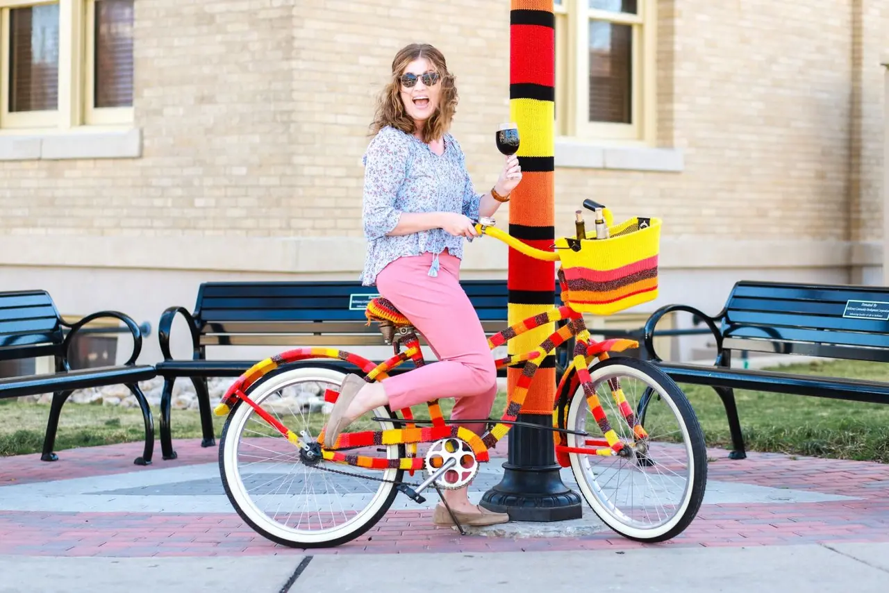 Lady wearing pink pants and a colorful blouse on a bike holding a glass of wine