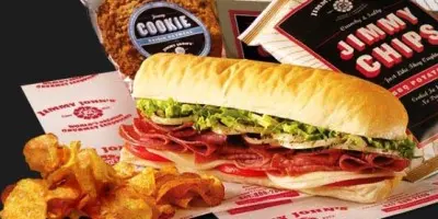 Submarine sandwich with sliced meat, lettuce, and tomato served with potato chips and branded packaging on dark background