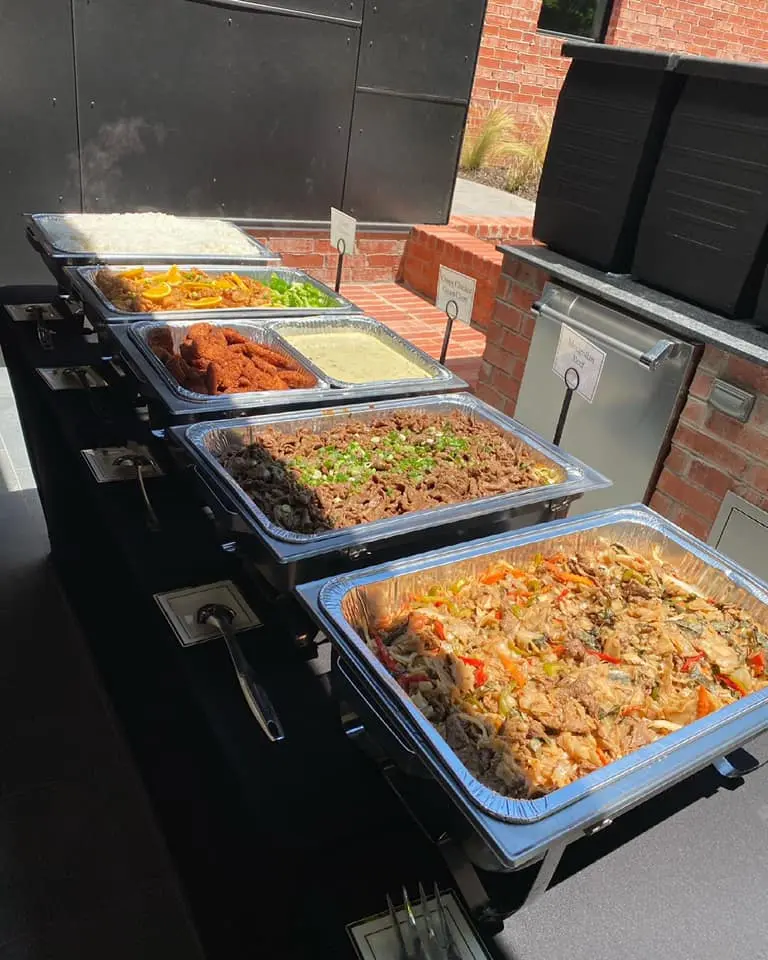 Multiple metal trays of prepared Thai dishes including noodles, curry, and stir-fry displayed on a serving table against a brick building