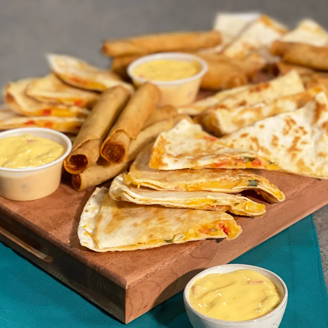 Crispy rolled tortillas and folded flatbreads with cheese and peppers, served with small cups of yellow dipping sauce on a wooden board