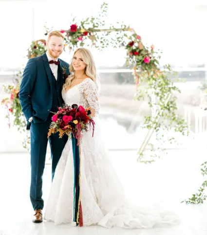 Bride and groom posing under a floral arch with red and gold flowers, bride holding cascading red bouquet