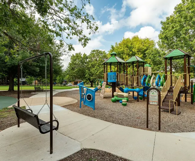 Playground with blue and wood climbing structures, swings, and slides surrounded by trees and open green space
