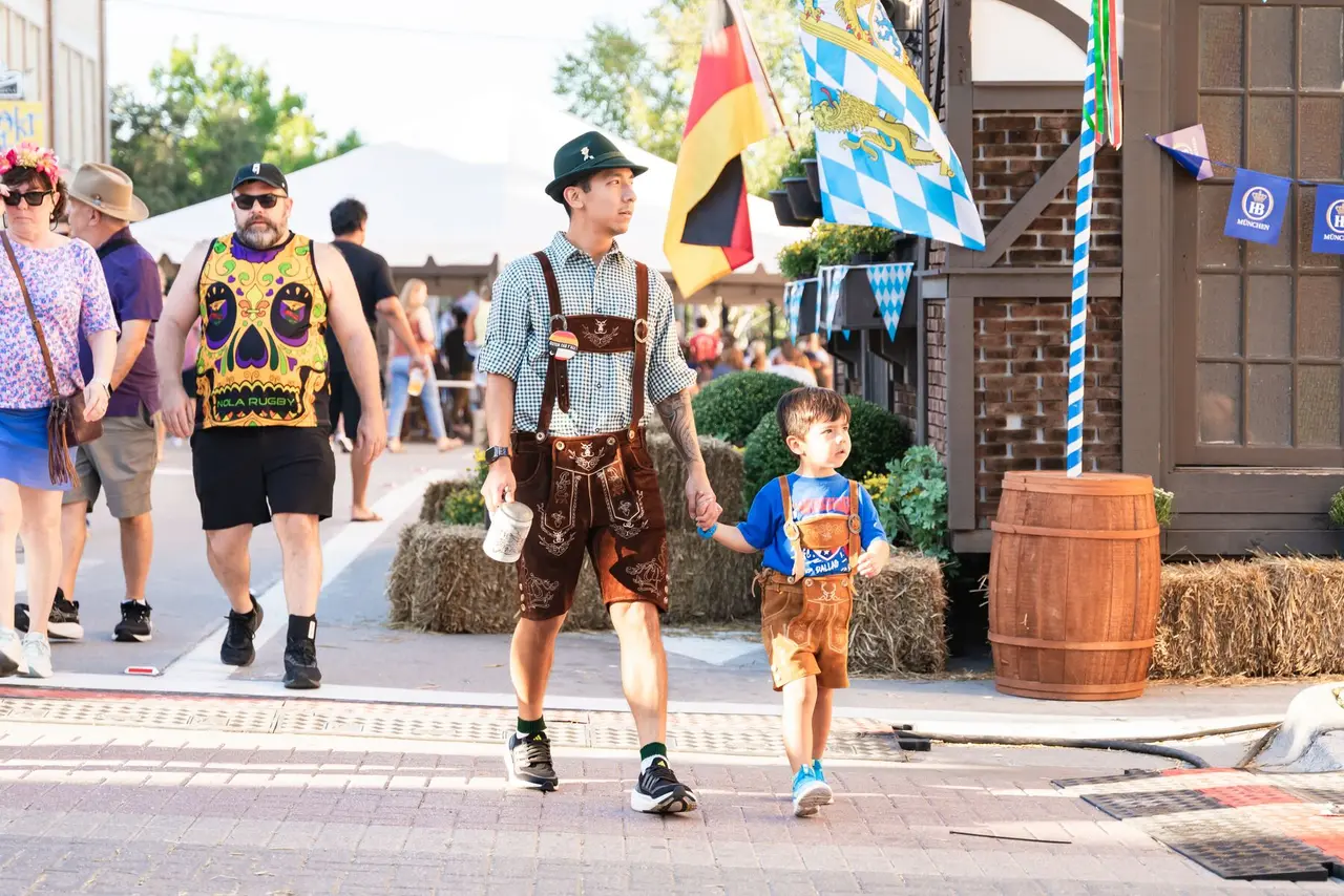 Adults and children in traditional Bavarian clothing walking past hay bales and wooden barrels, with flags and festival decorations in background