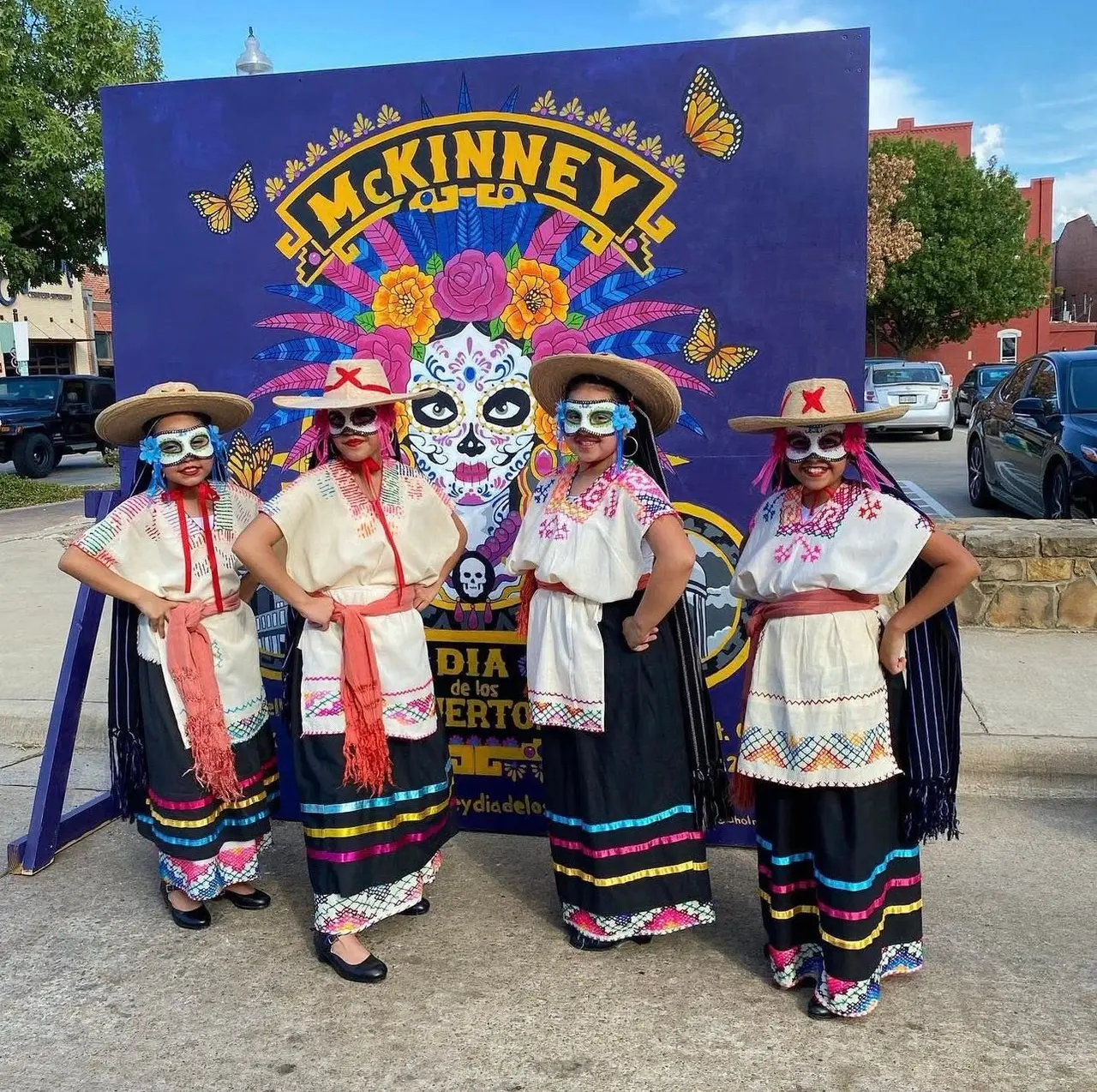 Four people in Día de los Muertos face paint and traditional colorful clothing standing in front of a large purple festival banner with skull imagery and