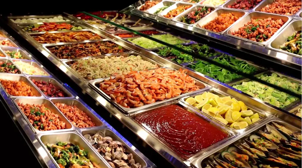 Multiple metal trays displaying colorful prepared Asian dishes including noodles, vegetables, proteins, and sauces in a buffet line