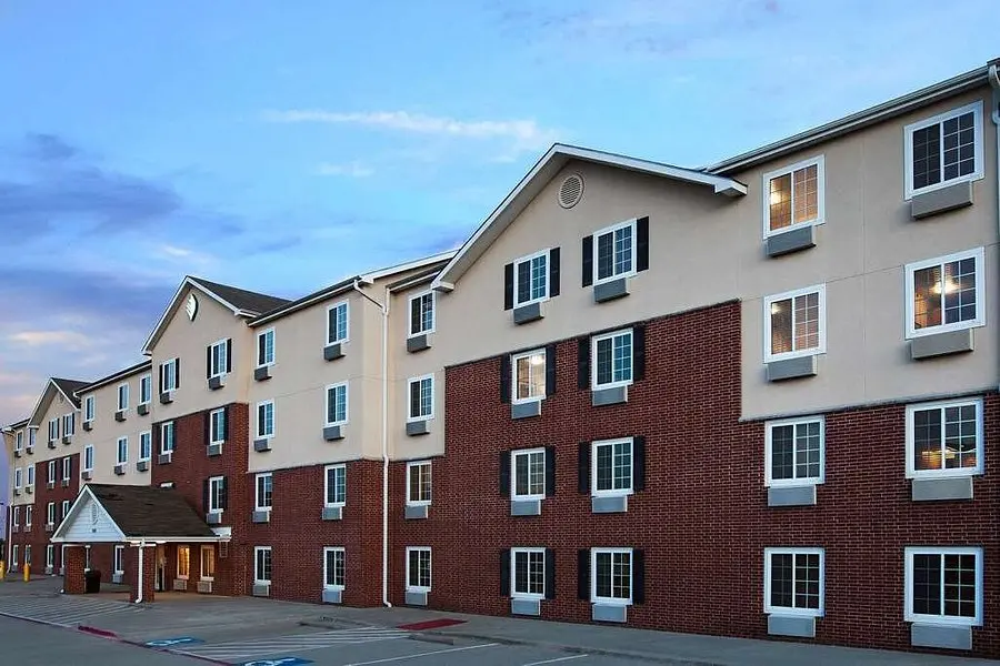 Multi-story hotel building with red brick and tan exterior, multiple windows with blue shutters, entrance area, and parking lot at dusk