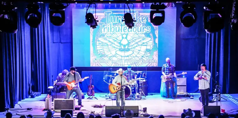 Band performing on stage with blue lighting and large screen backdrop displaying event artwork