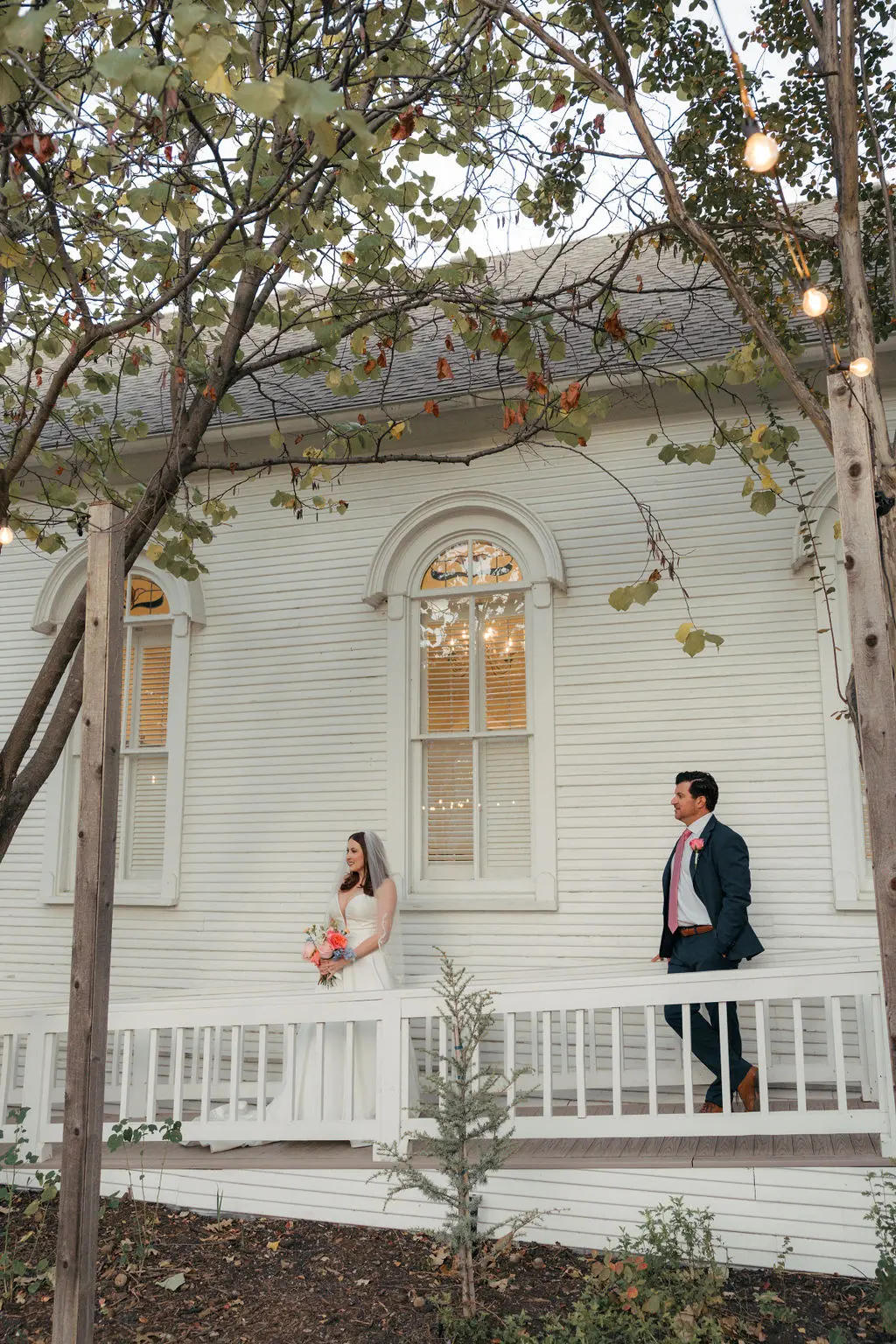 Bride and groom standing on the porch of a white wooden chapel with arched windows, framed by tree branches