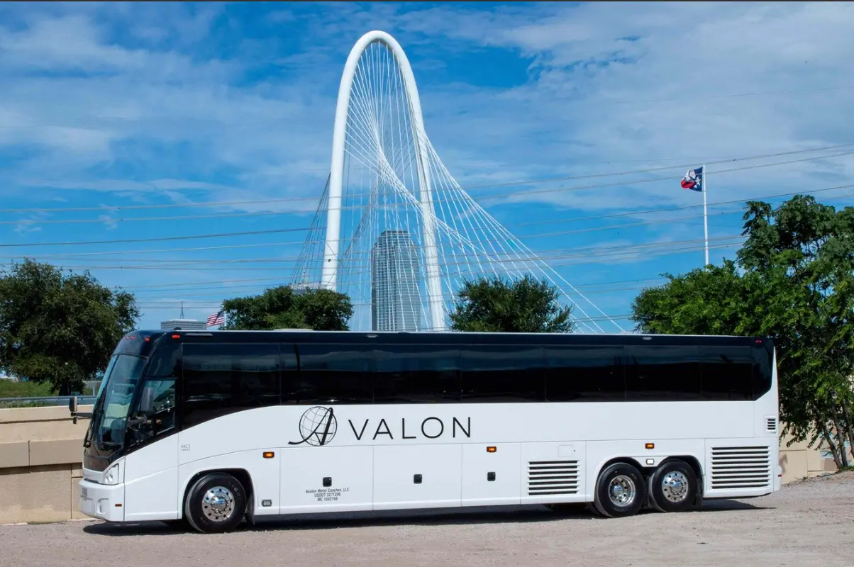 White tour bus parked in front of modern white arched bridge with blue sky and green trees