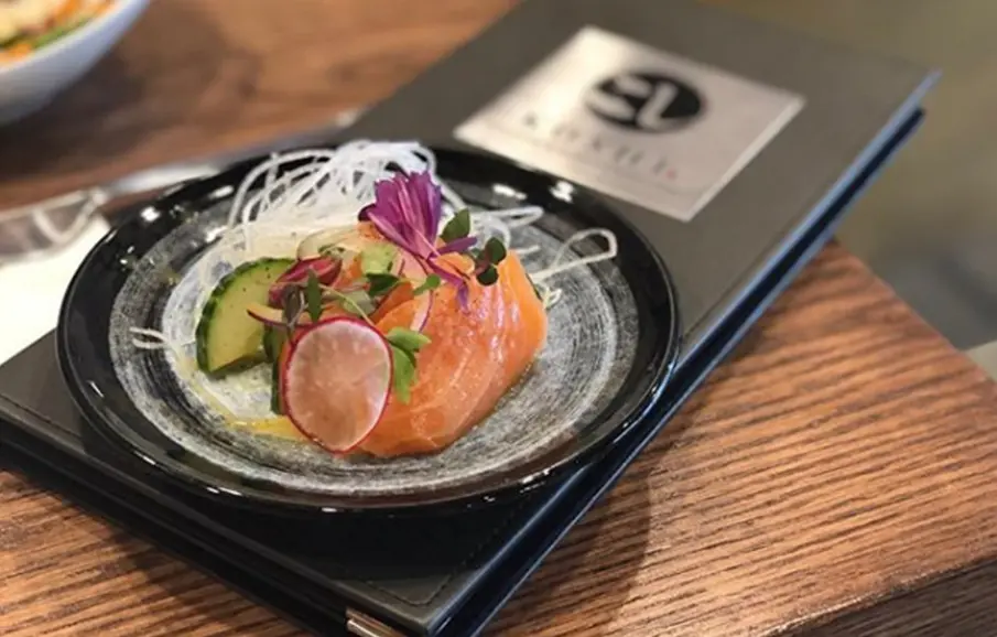 Sliced salmon and cured fish arranged on shredded daikon with purple flower garnish on black plate with chopsticks