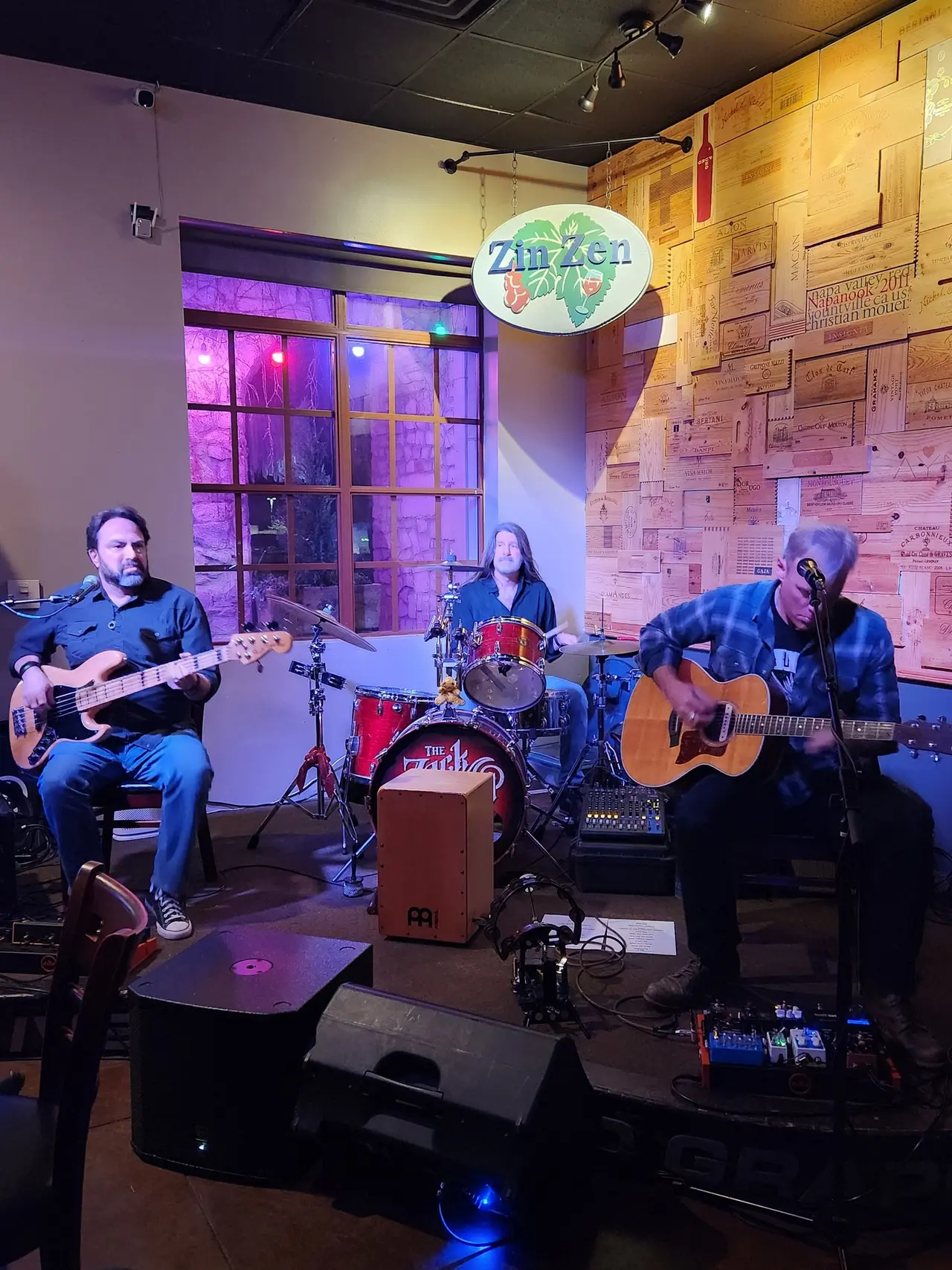 Three musicians performing with guitars and drums on a stage with purple lighting and wooden brick walls