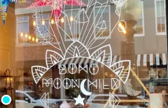 Boho-themed window display with white mandala and moon designs on glass, crystals and jewelry visible inside storefront